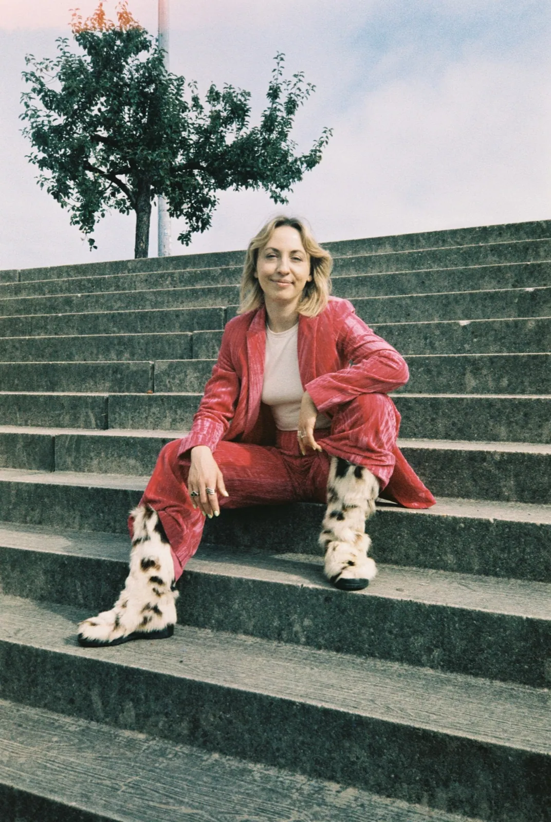 Luna Weis sitzt auf einer breiten Treppe aus Beton. Sie trägt einen pinken Anzug aus Samt und lacht.